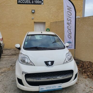 Peugeot 107 blanche d'occasion chez Detail Auto à Pamiers avec drapeau Occasion et panneau Accueil à 100m.