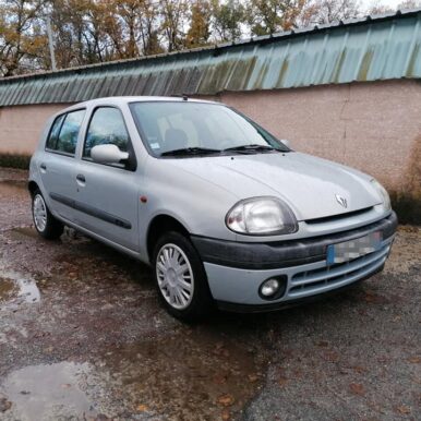 Renault Clio 2 grise 5 portes garée en extérieur sur un sol mouillé devant un mur.