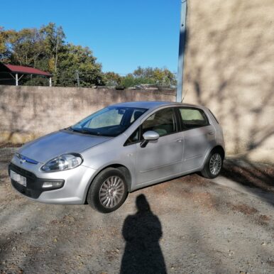 Fiat Punto grise stationnée sur un parking en gravier devant un bâtiment beige, sous un ciel bleu d'automne.